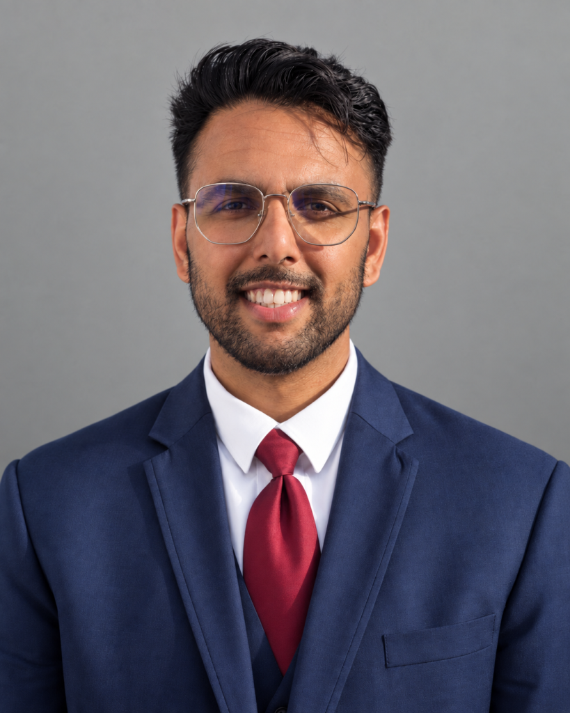 A smiling man in a blue suit, white shirt, red tie, and glasses stands before a plain gray background.