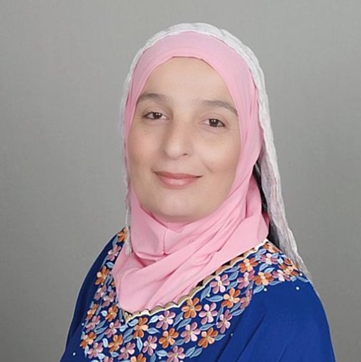 Woman wears a pink hijab and blue floral-embroidered top, smiling softly against a gray background.